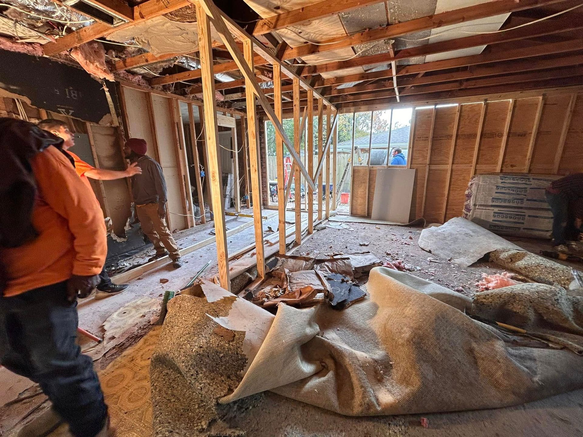 Interior demolition in progress, showing exposed framing and debris. Construction workers are present, removing walls and materials.