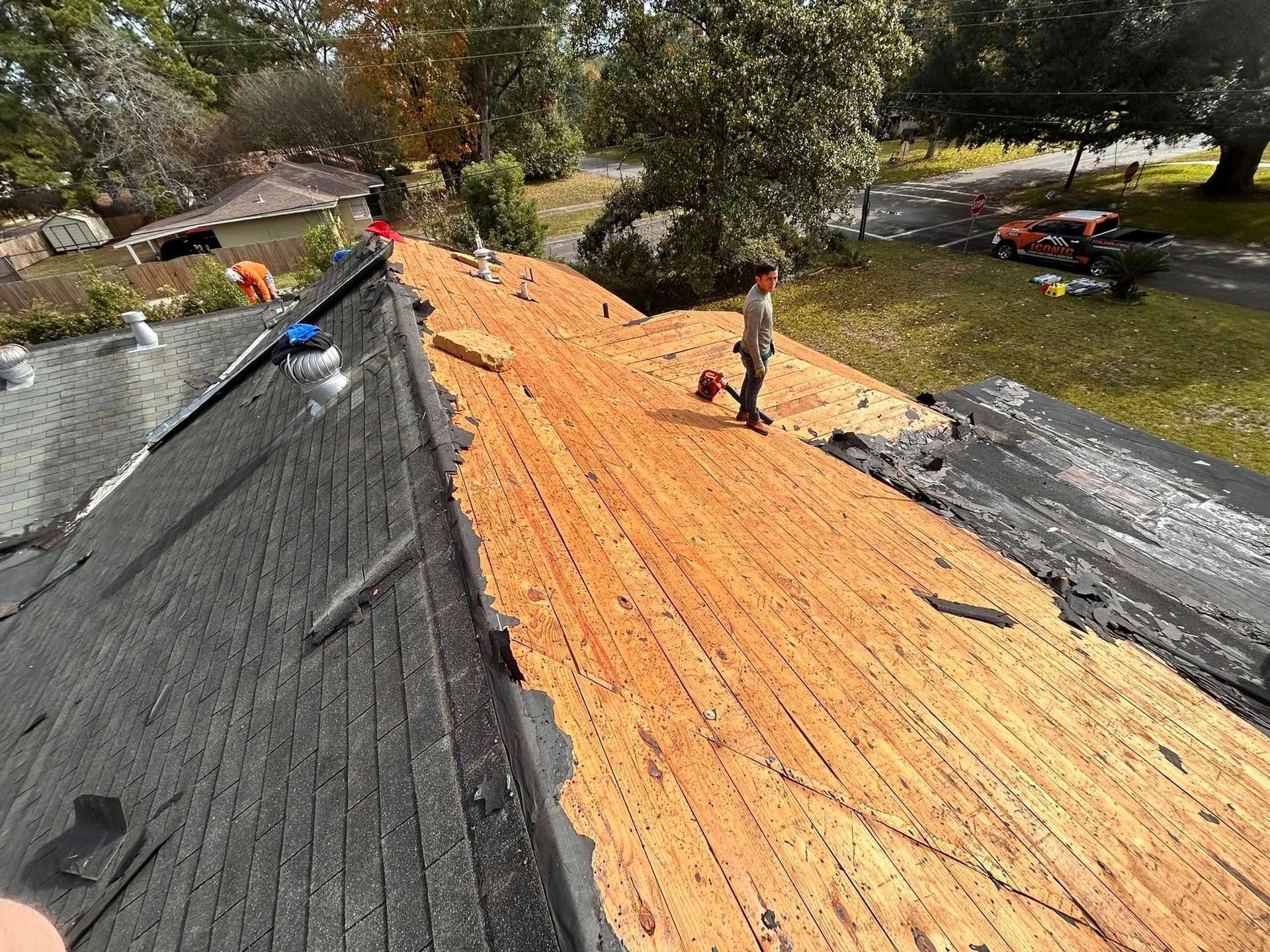 Roofers remove old shingles from a house roof. One worker stands holding a tool, another is visible in the distance.