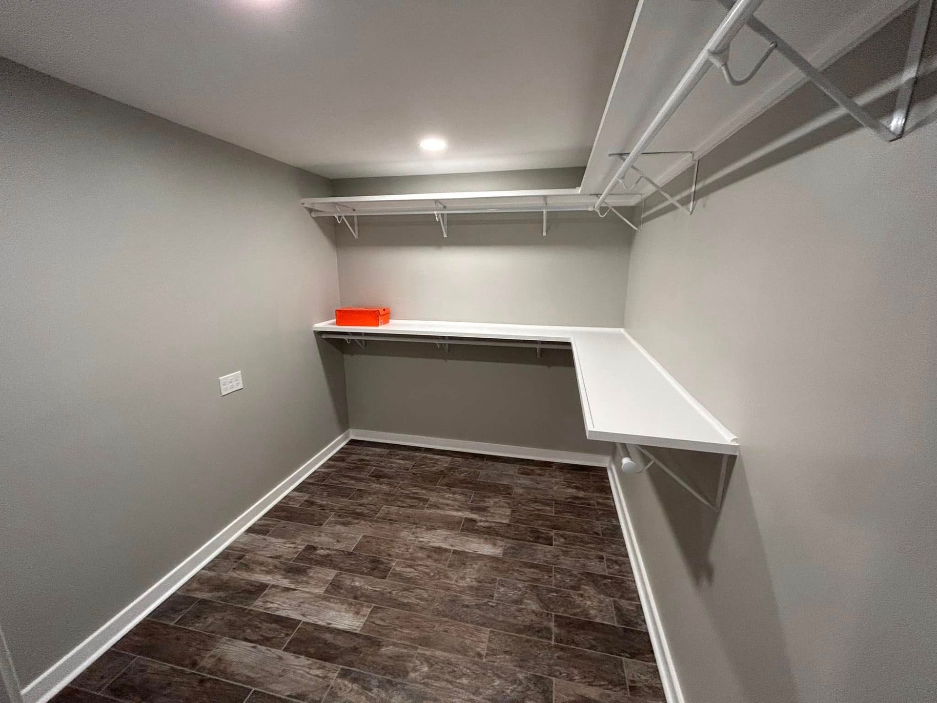 Empty walk-in closet with gray walls, dark wood-look floor, and white shelving. An orange box sits on a shelf.