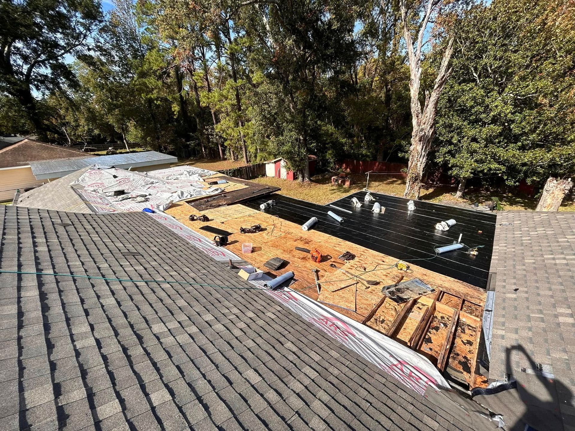 Roof under construction with exposed wood and tarps, surrounded by trees.