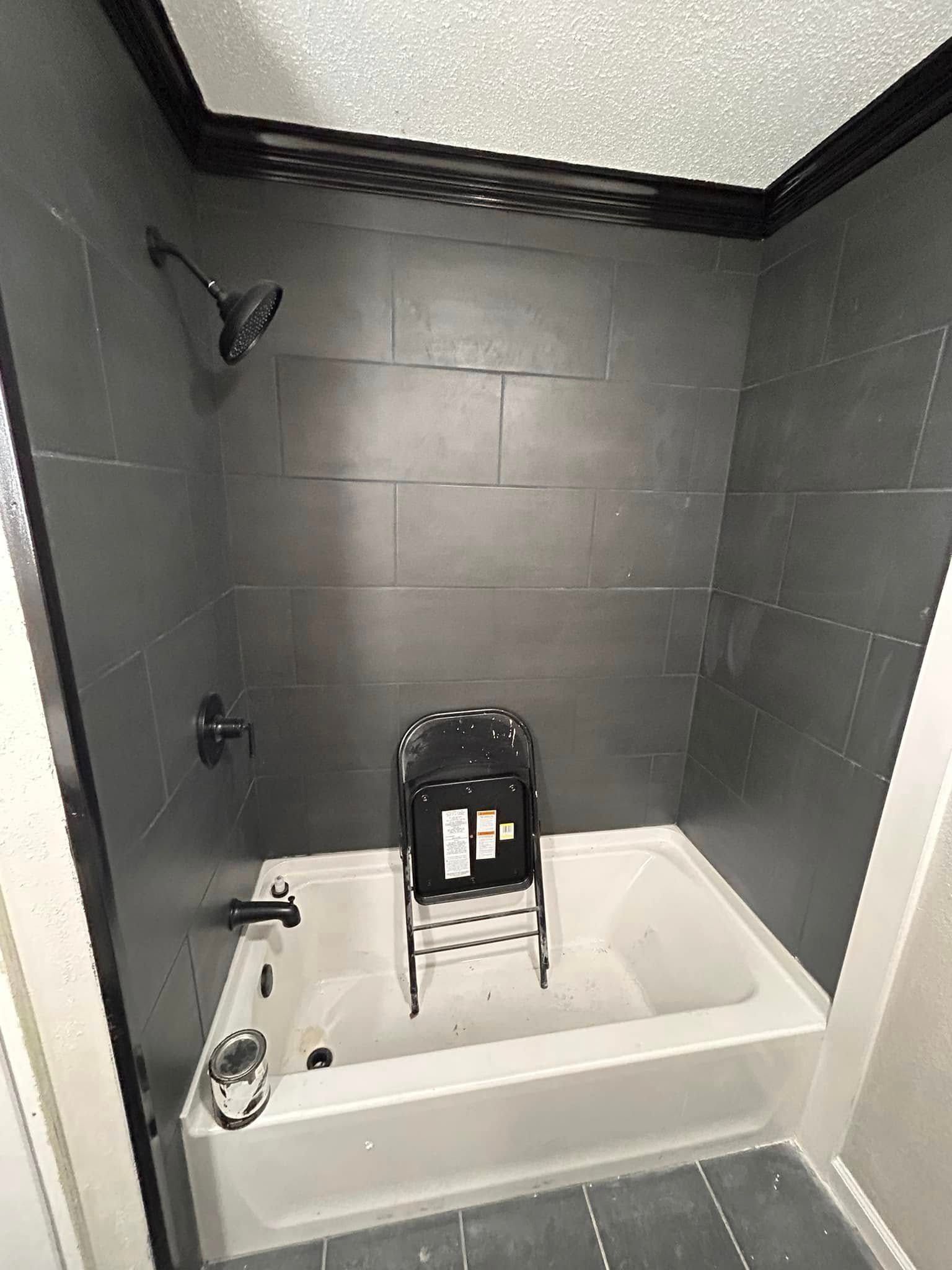 A shower with dark gray tiled walls and a white bathtub. A black shower chair sits inside the tub.