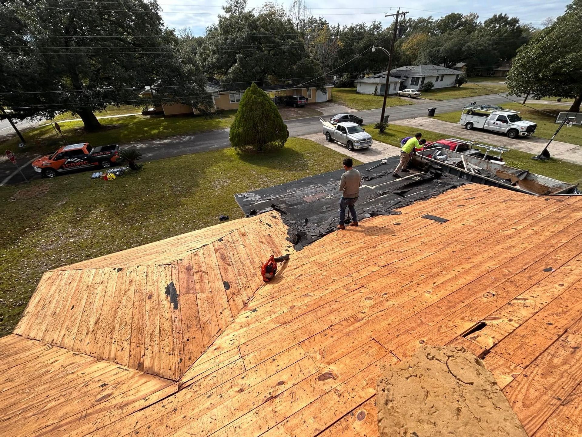 Roofers removing shingles from a house, with work trucks and other houses in the background.