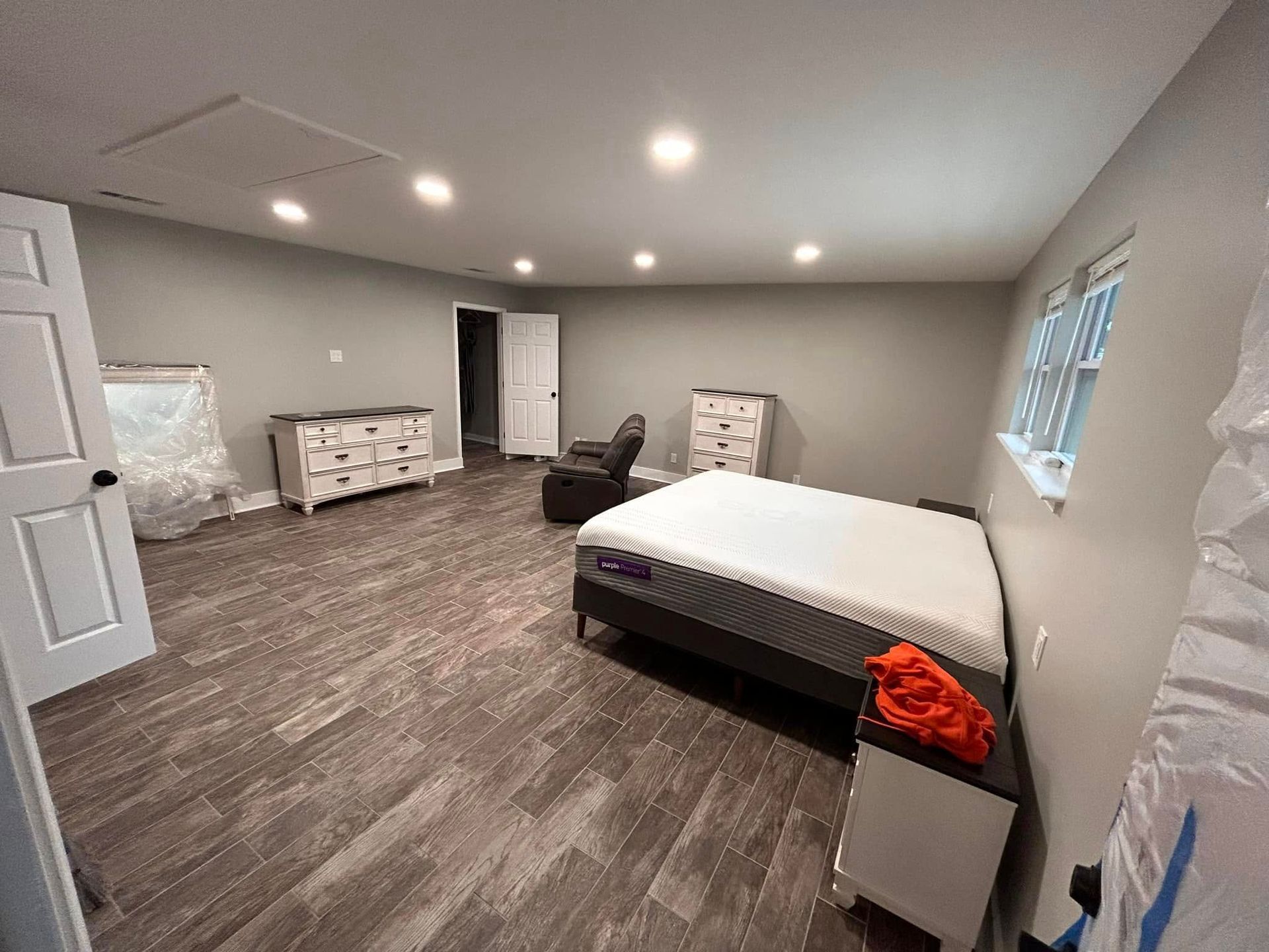 A spacious bedroom with grey walls, wood-look flooring, and a mattress on a bed frame. There's a recliner, dressers, and a nightstand present.