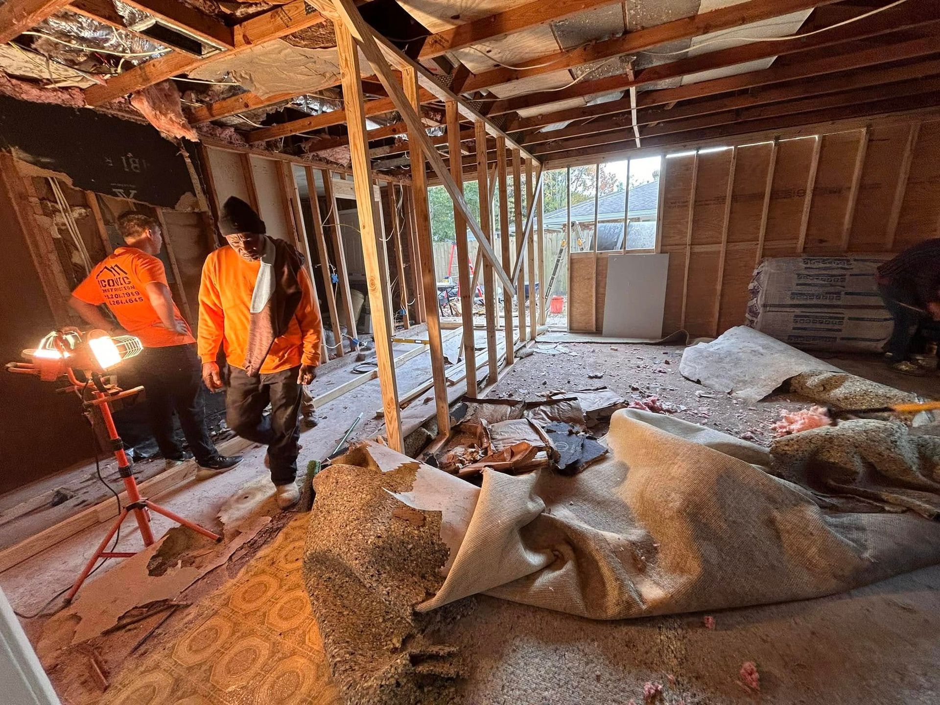 People in orange vests renovate a room with exposed framing and torn carpet. Bright work light illuminates the debris.
