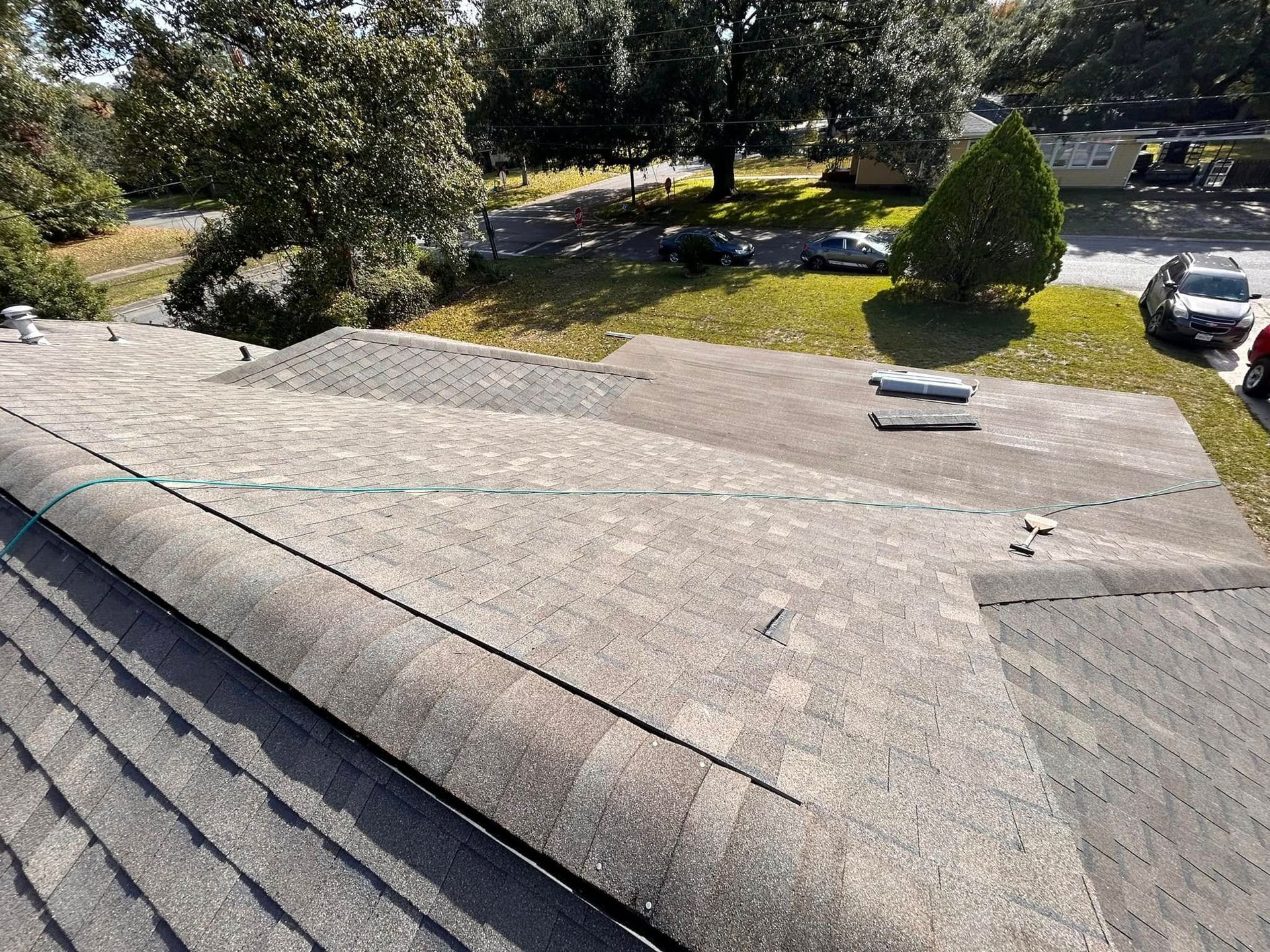 A close-up of a gray shingled roof with a safety line, overlooking a grassy area, trees, and parked cars.