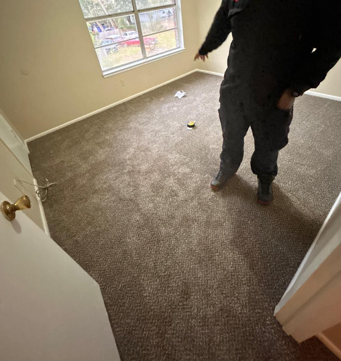 Person stands in a room with brown carpet, near a window and open door. The walls are tan.