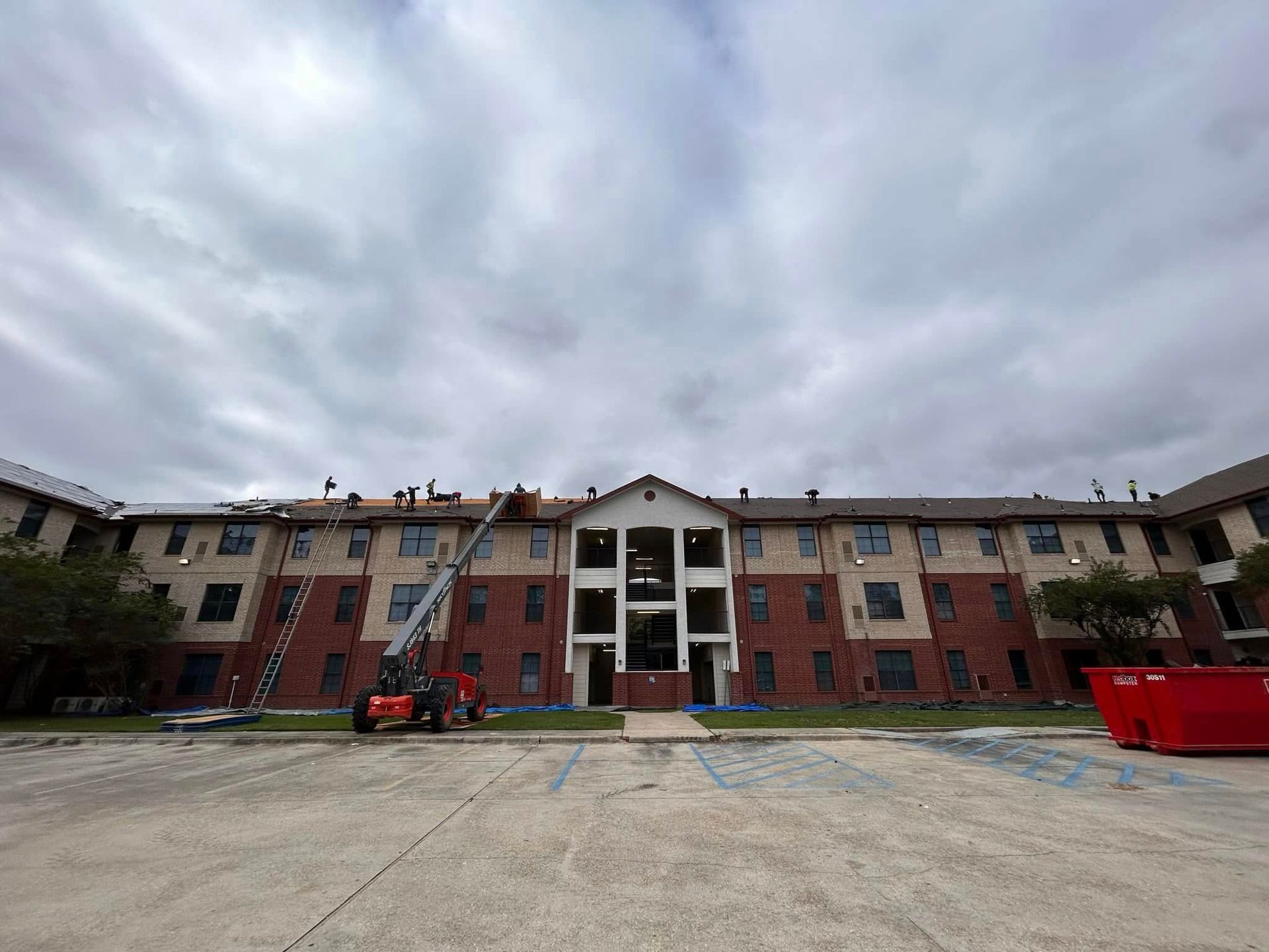 Construction workers on a building roof under a cloudy sky. A lift and debris container are in the foreground.