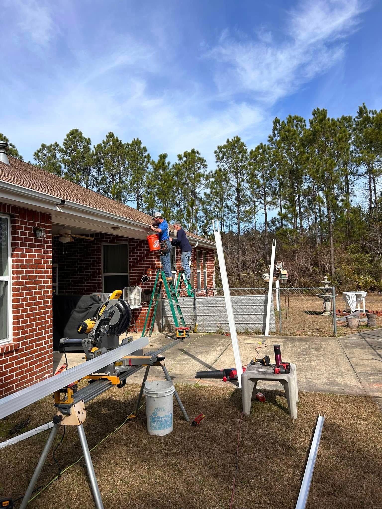 Two people work on a roof, using ladders and tools. A brick house is visible on a sunny day.