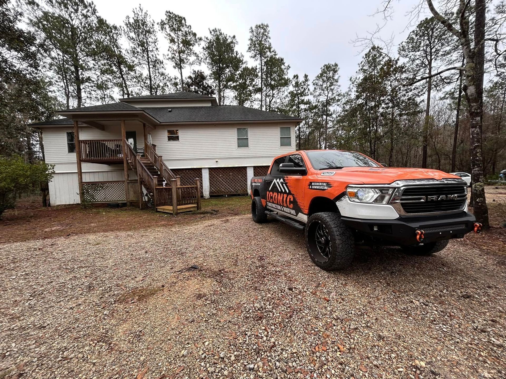 Orange pickup truck parked in front of a weathered white house. The setting is a gravel driveway surrounded by trees on an overcast day.