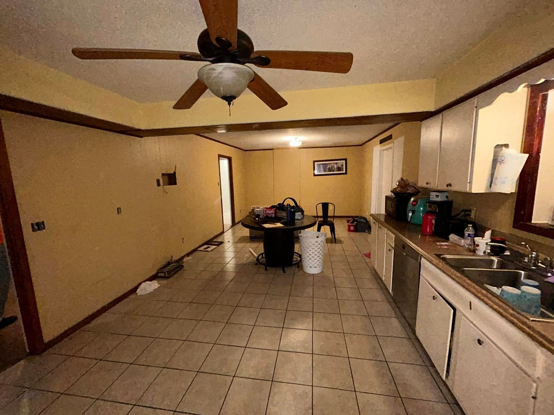 Long, narrow kitchen with tile floor, yellow walls, white cabinets, and a ceiling fan. A table and chair are in the middle of the room.
