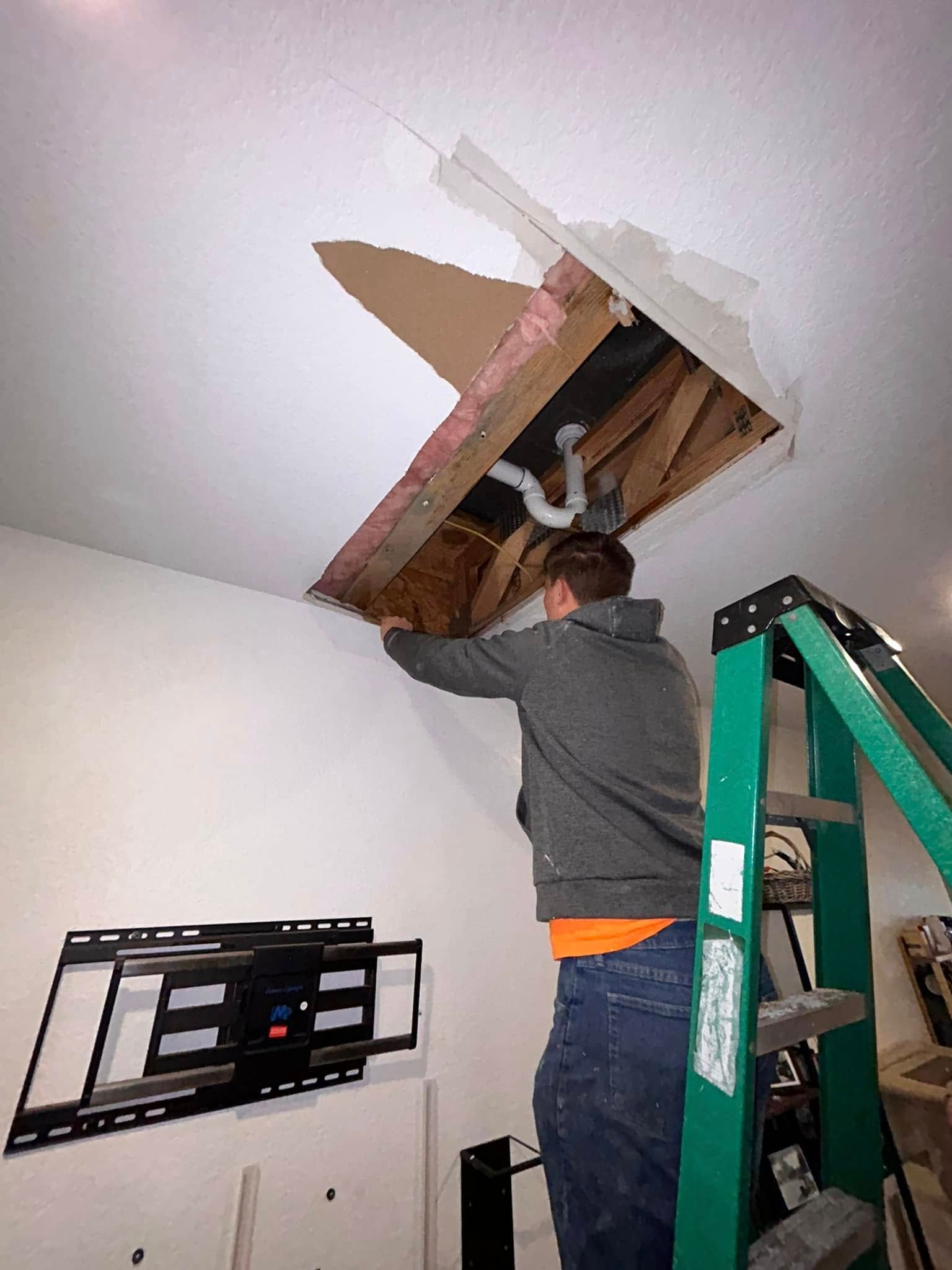 Man repairing ceiling damage, standing on a green ladder. He's looking up at the exposed plumbing and wood. A TV wall mount is on the left.