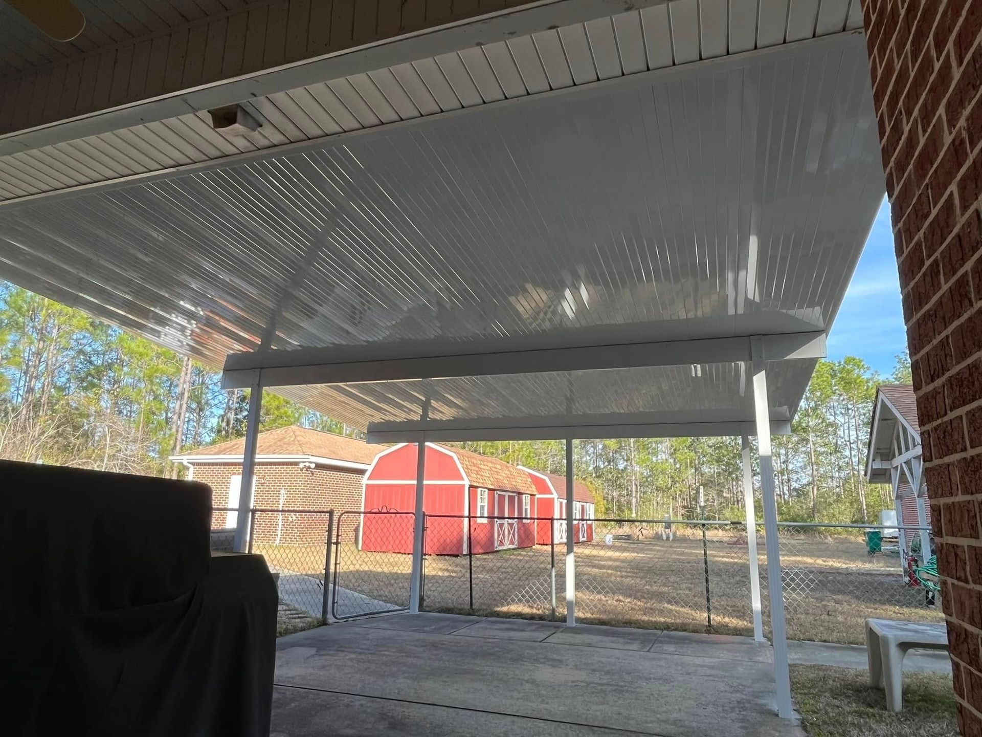A white-roofed carport attached to a brick house with a view of a red barn and fence in the background.
