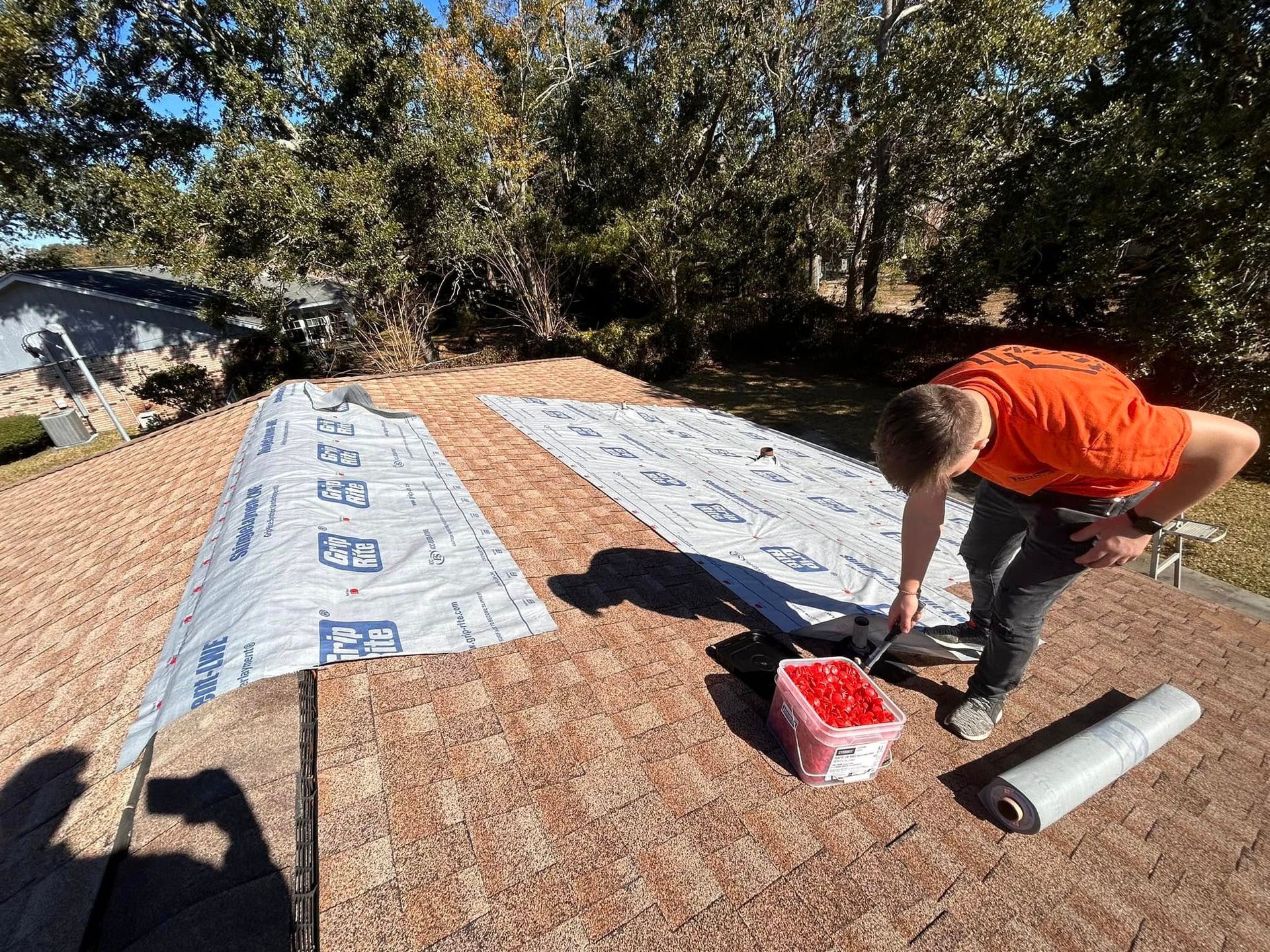 A worker on a rooftop installs roofing material on a sunny day, holding a tool near a roll and container of supplies.