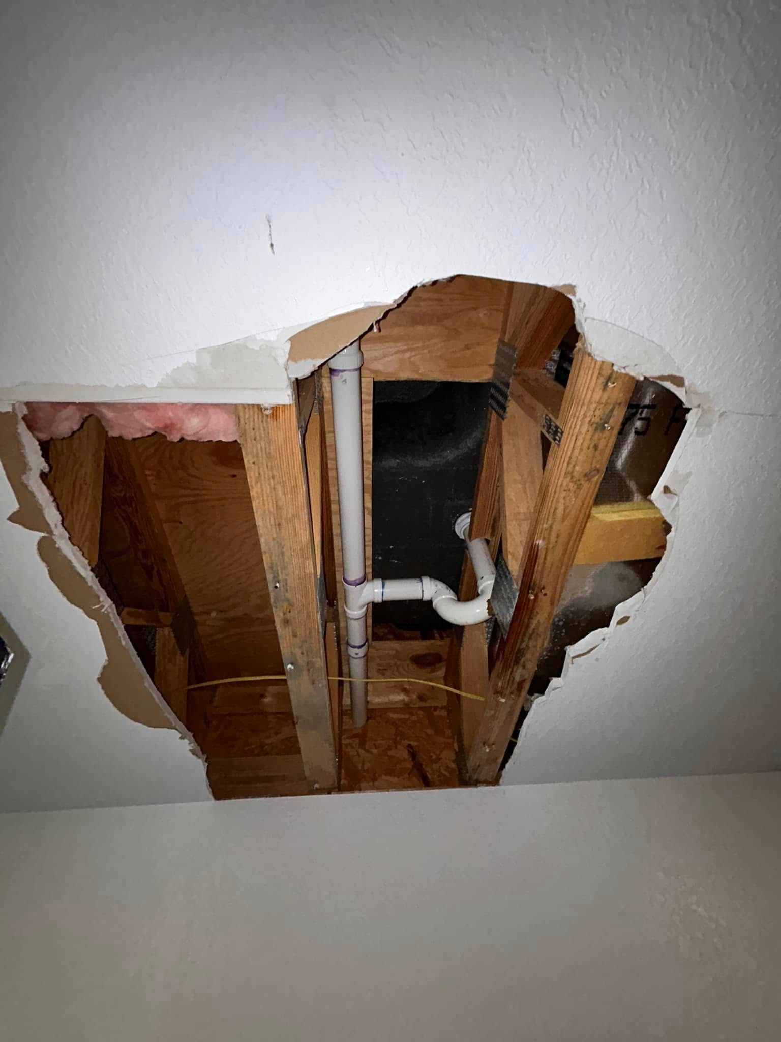 Hole in a white ceiling revealing wooden beams and a white plumbing pipe. The ceiling is damaged around the opening.