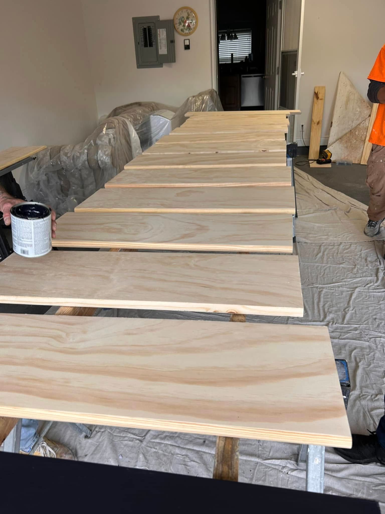 Person painting wooden planks laid out on sawhorse tables in a garage.