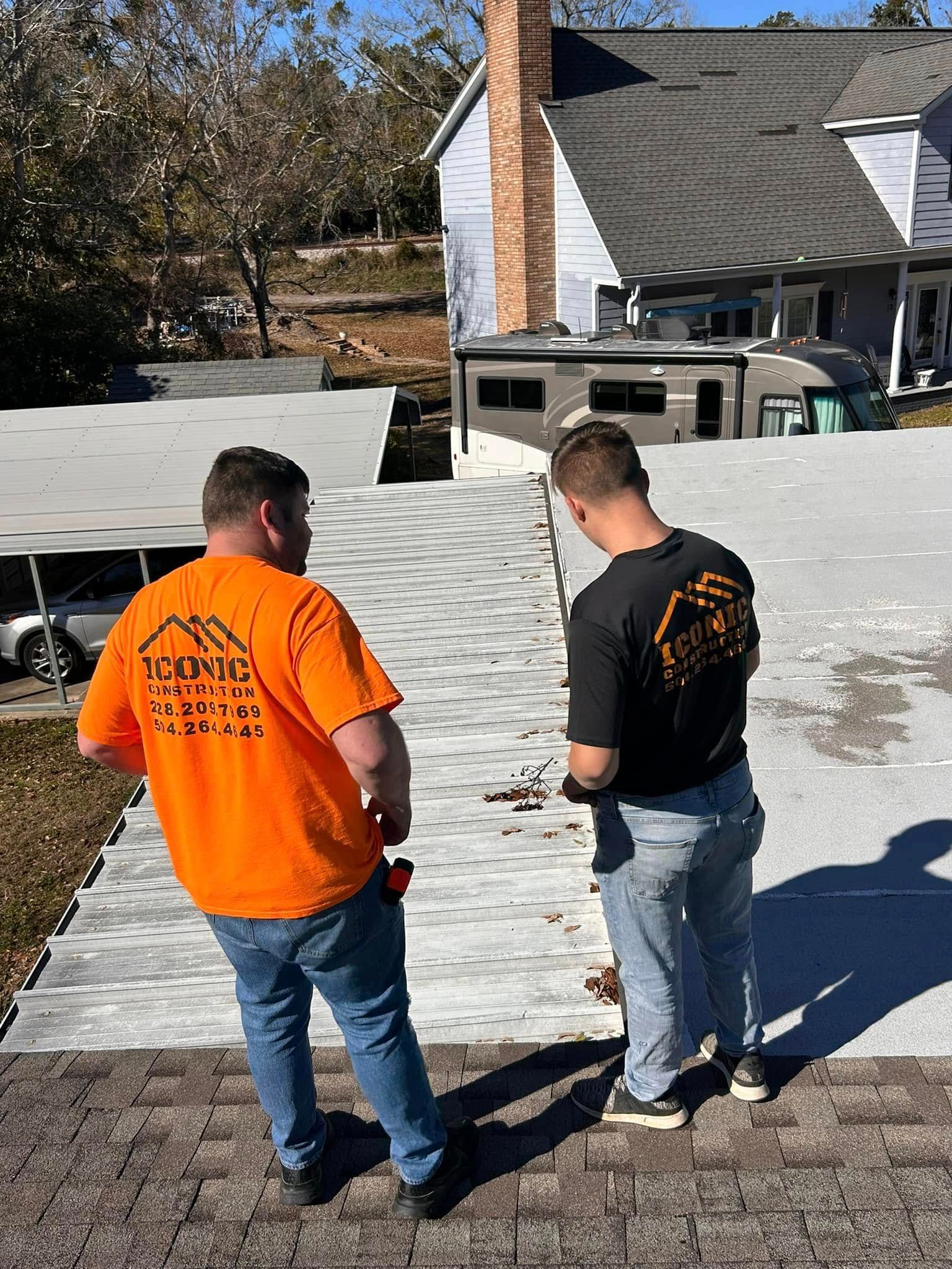 Two men, one in orange, the other in black, stand on a roof inspecting shingles. The sky is clear, and a house is in the background.