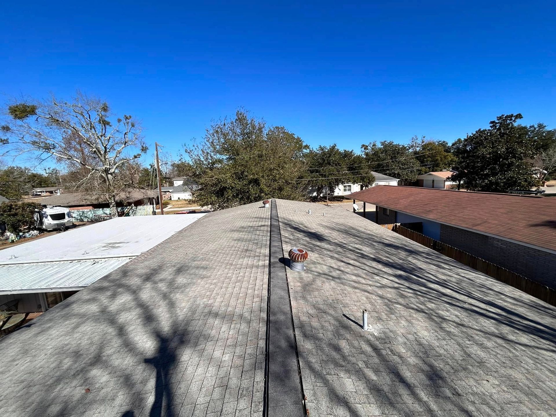 View from a rooftop with weathered grey shingles. A clear blue sky is above with trees and surrounding houses in the distance.