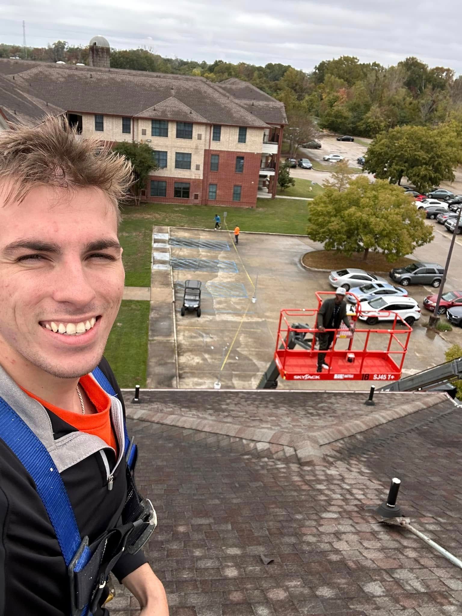 Man on a rooftop smiling, taking a selfie. Workers on a red lift are in the background, in front of an apartment building and a parking lot.
