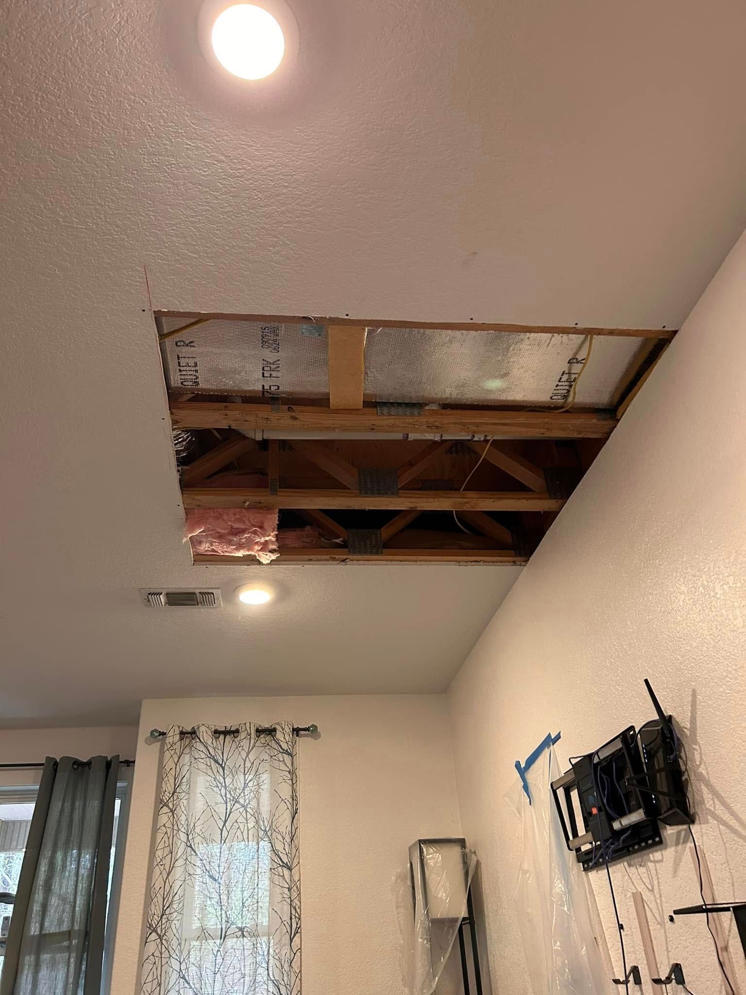 Hole cut in a white ceiling, revealing wooden beams and insulation. A mounted TV bracket is on the adjacent wall.