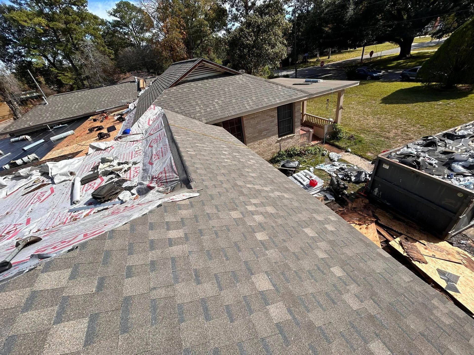 Roof of a house partially torn up for repair, with shingles and underlayment exposed. A dumpster sits nearby, with a yard and trees in the background.