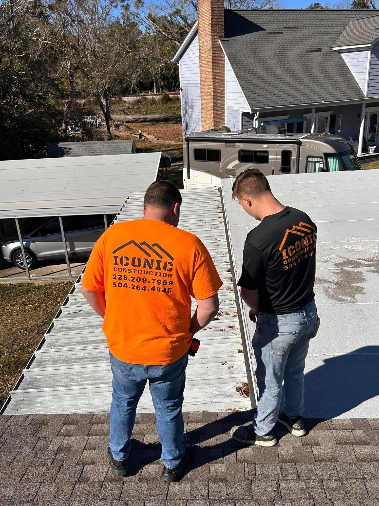 Two roofers in orange and black shirts on a rooftop. They are working on the roof shingles of a house on a sunny day.
