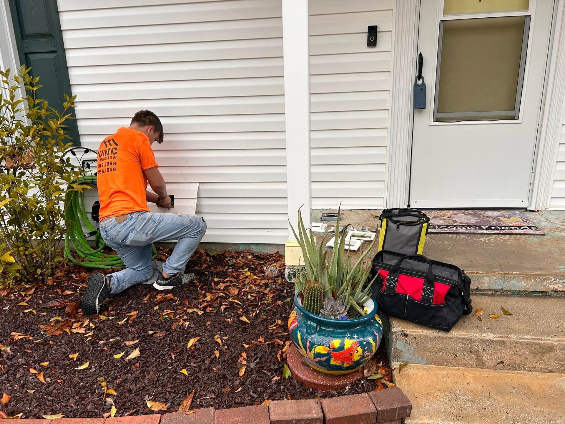 A man in an orange shirt kneels, working on the siding of a house. Tools and a potted plant sit nearby on the porch.
