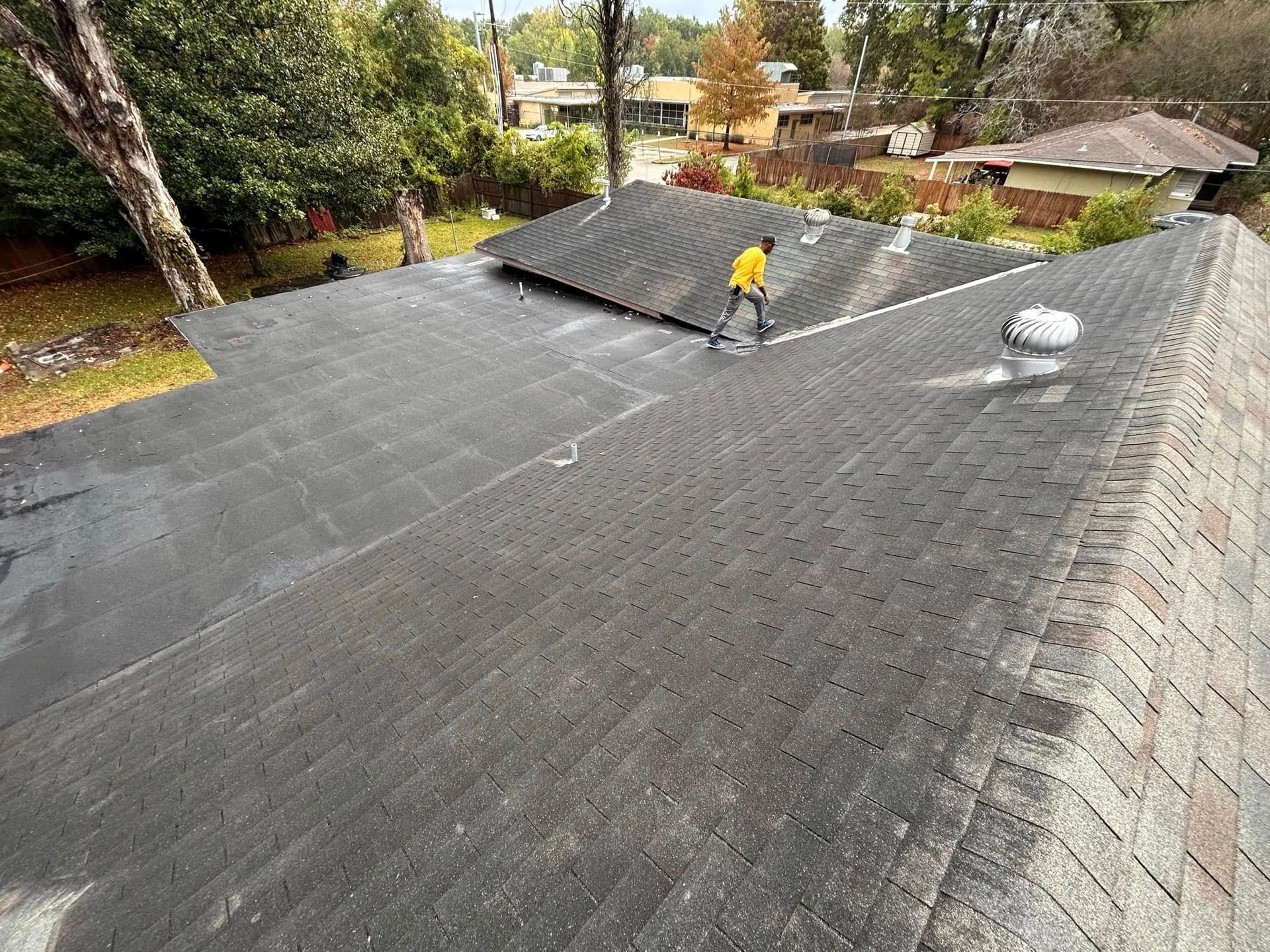 Roofer in yellow shirt repairing dark asphalt shingle roof from an aerial view, surrounded by trees and other houses.
