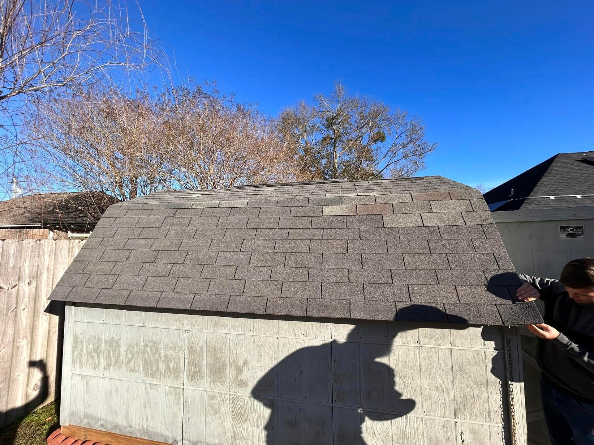 A person inspects the roof of a gray cinder block shed under a clear blue sky. The roof is covered in gray shingles.