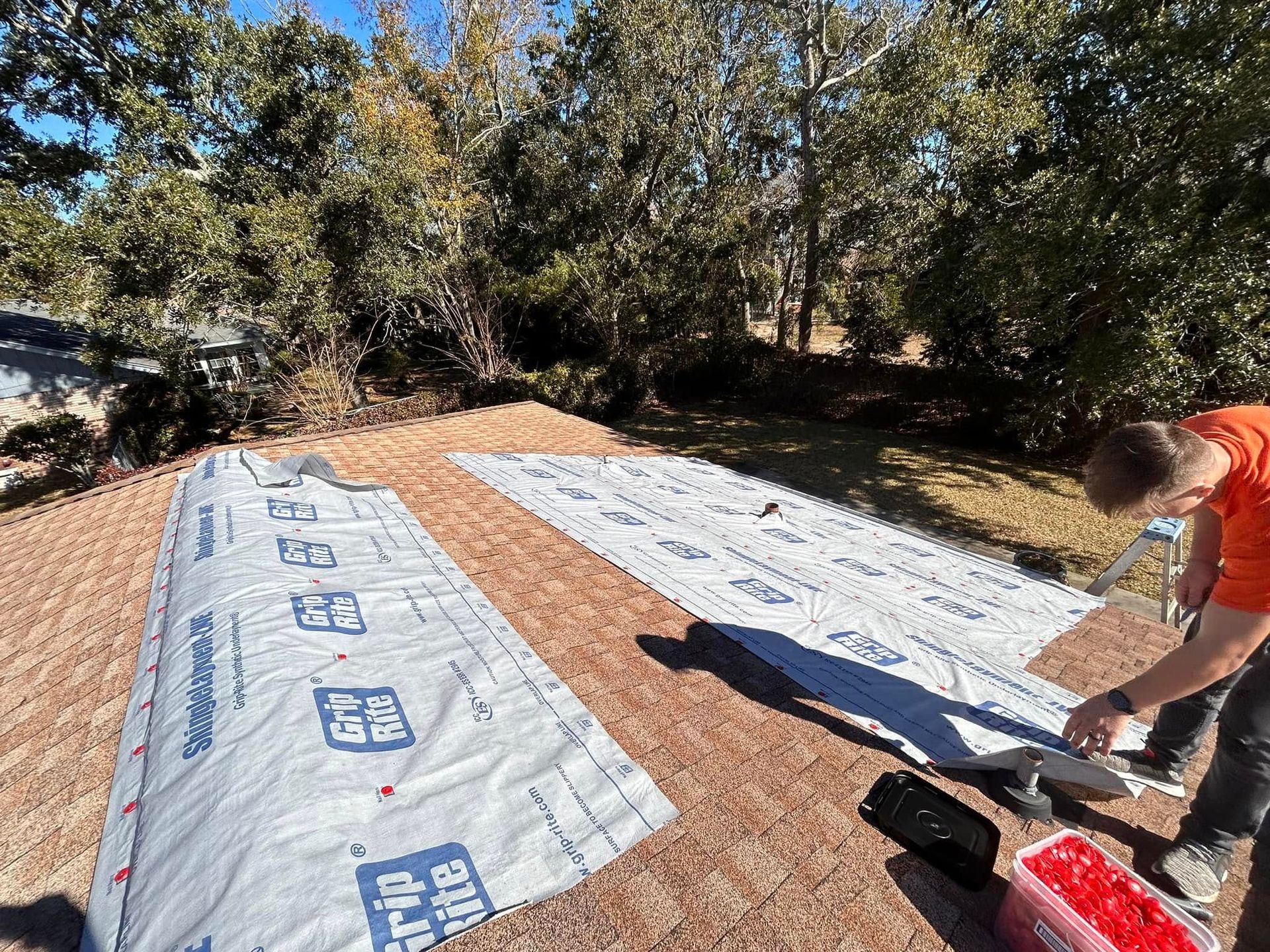 A worker on a roof installing underlayment; a roll is visible. Green trees and blue sky in the background.