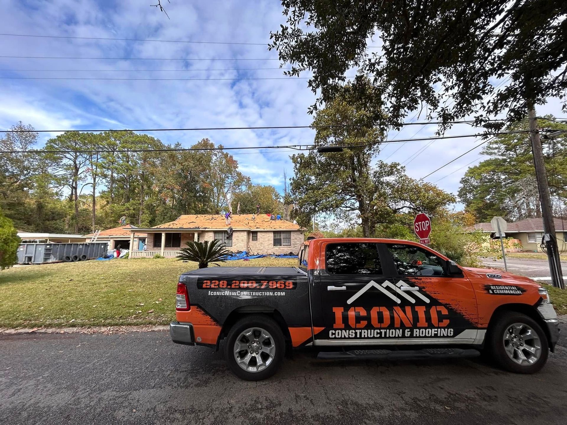Orange and black Iconic Construction truck parked in front of a house with roofing in progress. A cloudy sky is above.