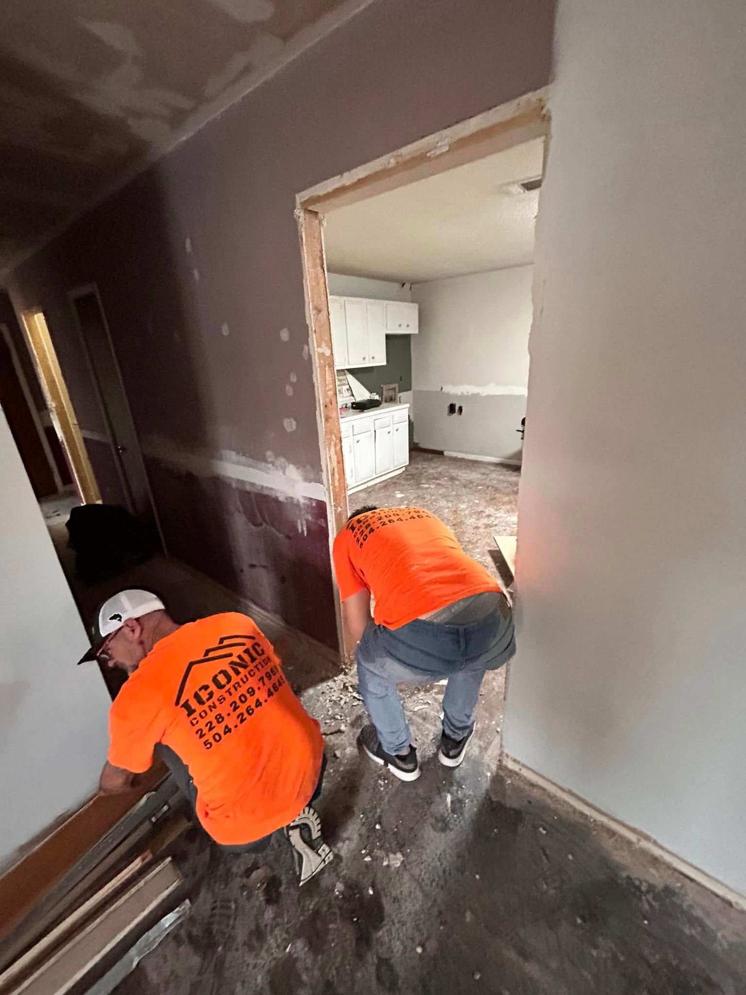 Two construction workers in orange shirts are installing trim in a doorway, inside a room under renovation.