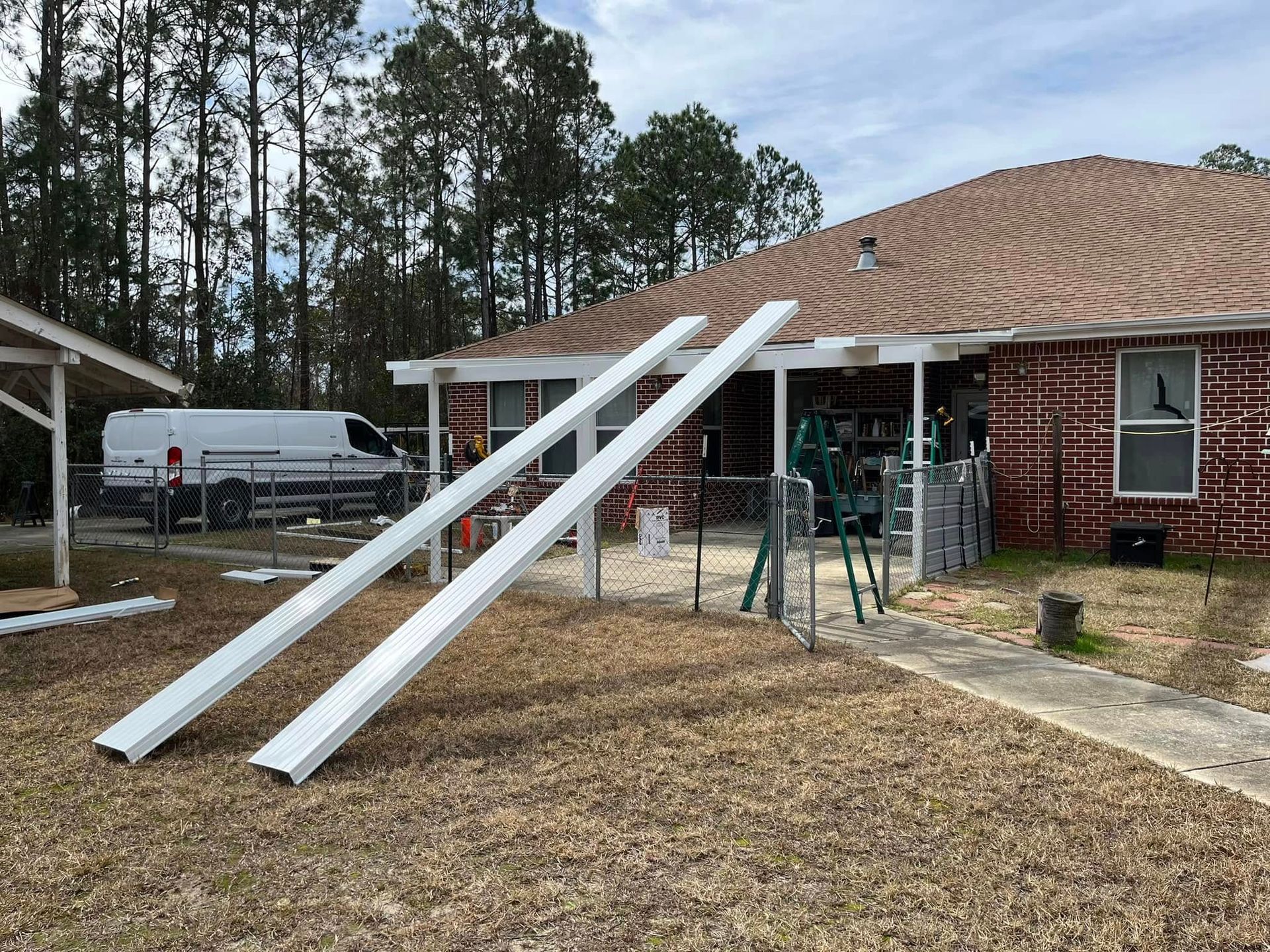 Two white metal beams lie on the brown grass in front of a brick house with a brown roof. A white van and construction equipment are visible.