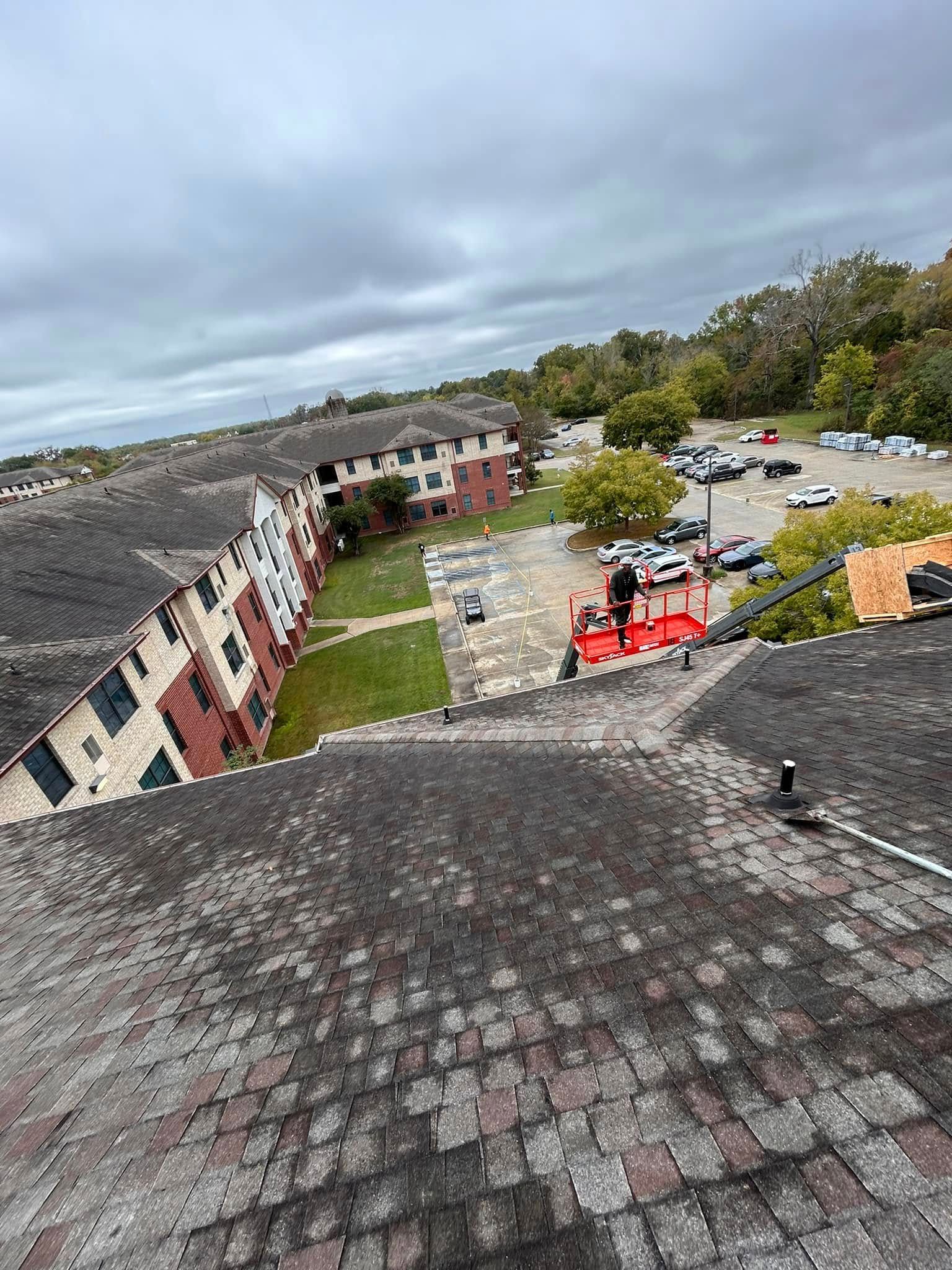 View of a building roof with visible damage and a worker on a lift; other buildings and vehicles are visible in the background on an overcast day.