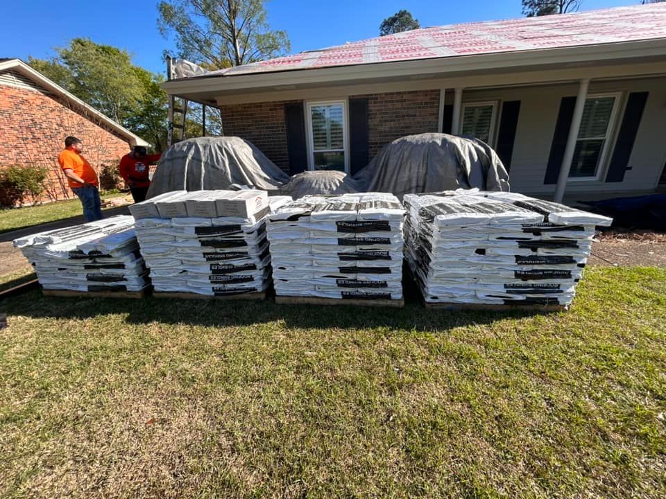 Stacks of new gray roof tiles on pallets in front of a house under construction. Two workers stand nearby.