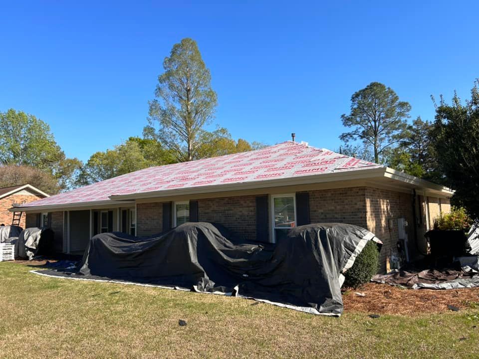 A one-story brick house with a roof in progress. Tarps cover the ground. Trees and a clear blue sky are in the background.