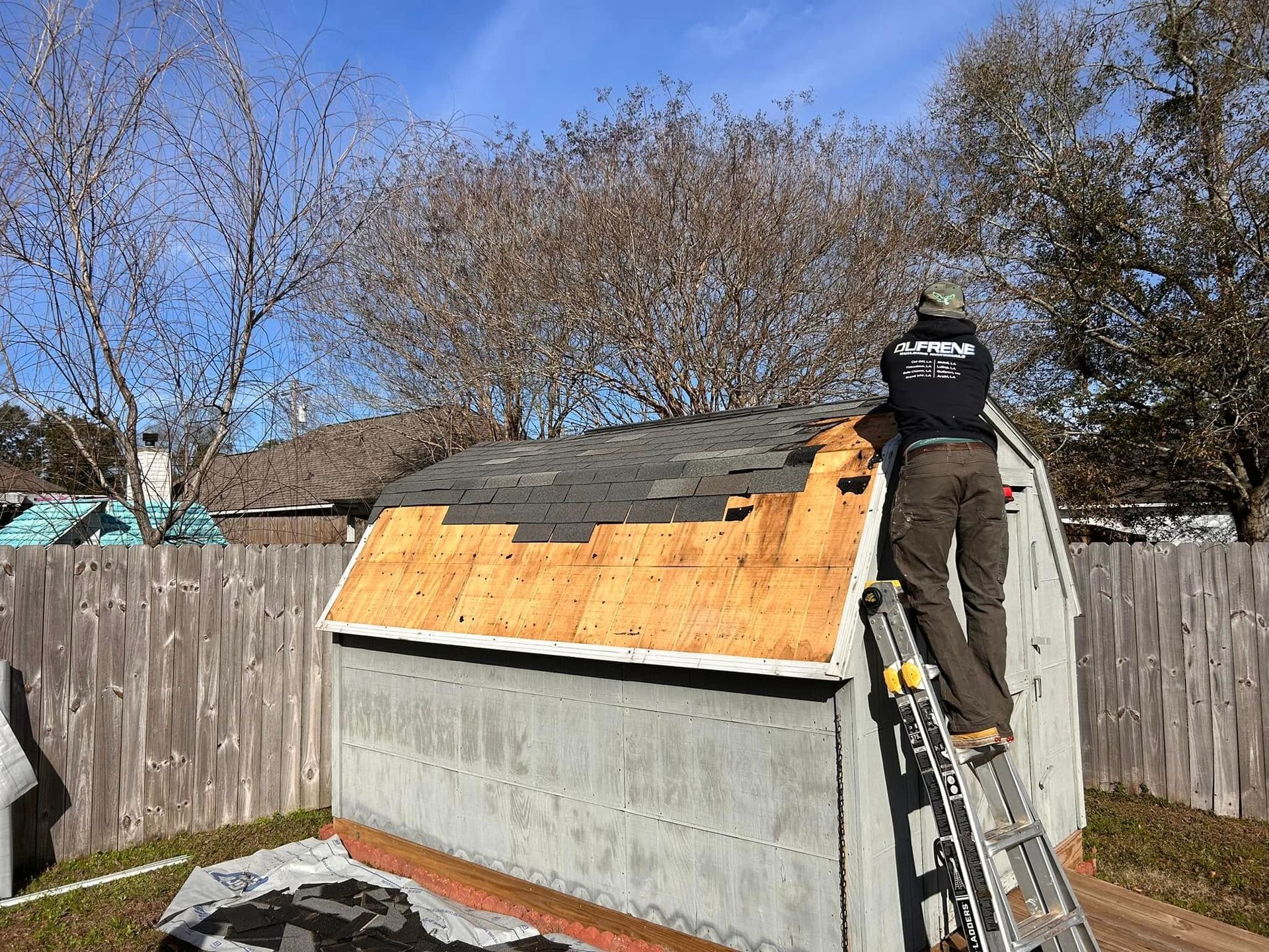 Person on a ladder, repairing a shed roof by removing old shingles. Bright sunny day, backyard setting.