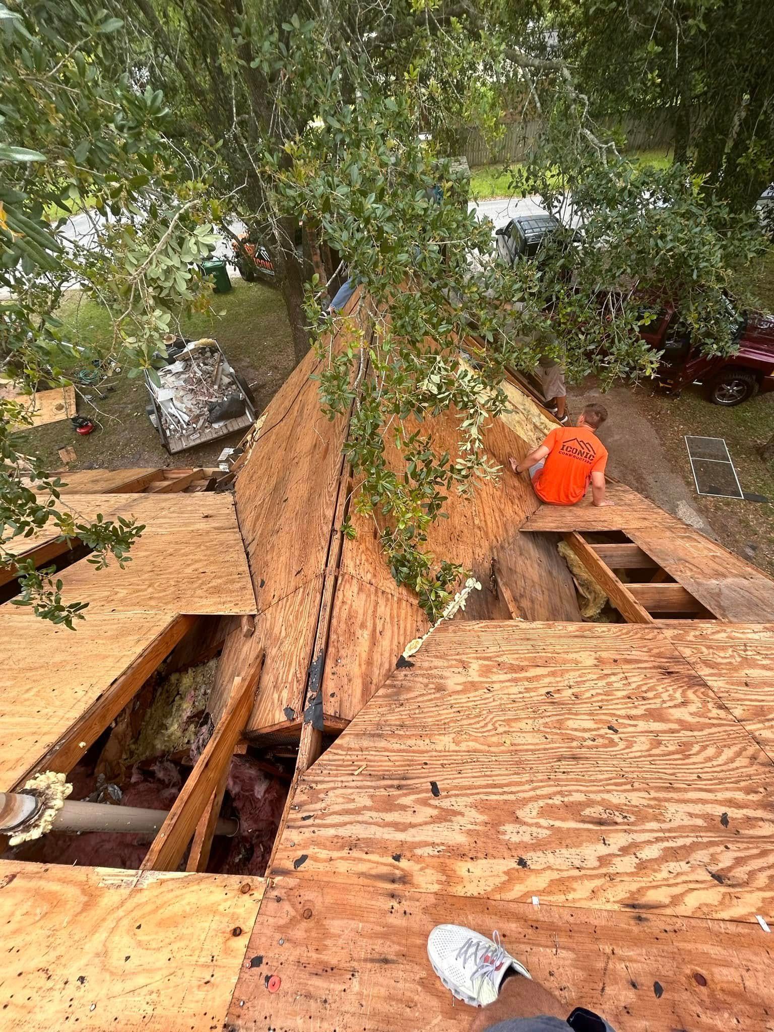 Roof under construction with exposed plywood, an open hole, an orange bucket, and a person's foot. Green trees are visible in the background.