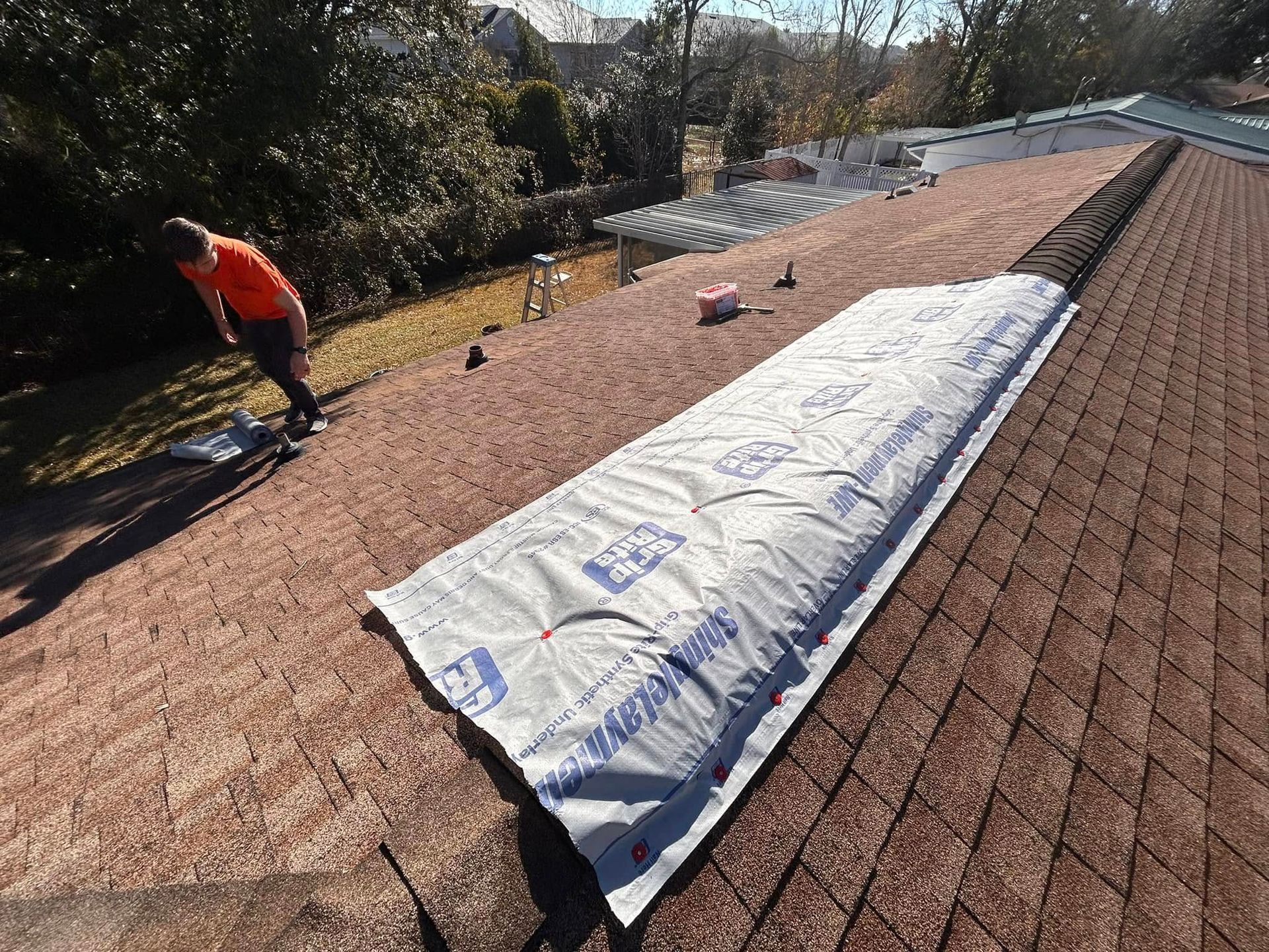 Roofer on a shingled roof, installing a rolled underlayment. Sunny outdoor setting with a residential backdrop.