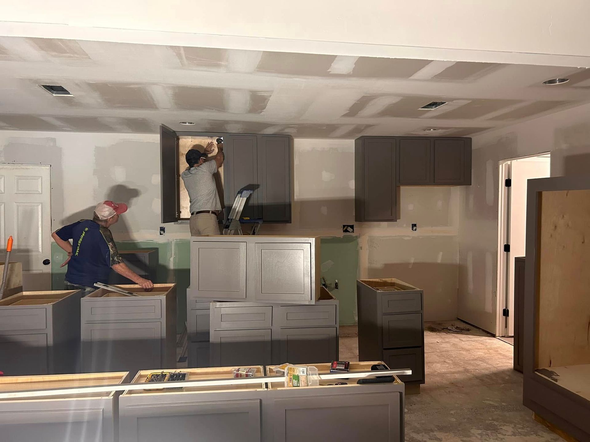 Two workers installing kitchen cabinets in a room under renovation. One is on a cabinet, the other is measuring a board.