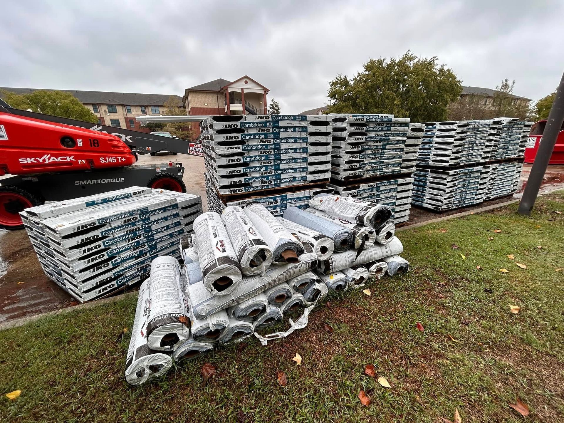 Piles of roofing materials, including shingles and rolled tar paper, staged outdoors near an apartment complex, ready for installation.