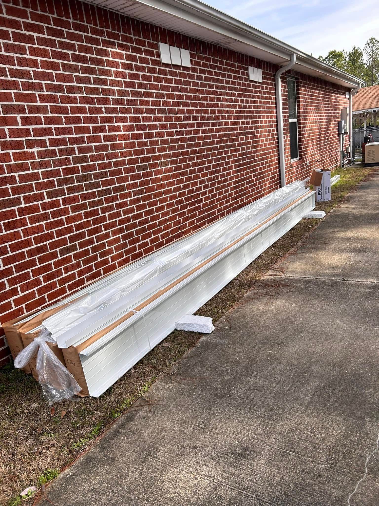 Long, silver aluminum beams wrapped in plastic are leaning against a red brick building on an asphalt surface.