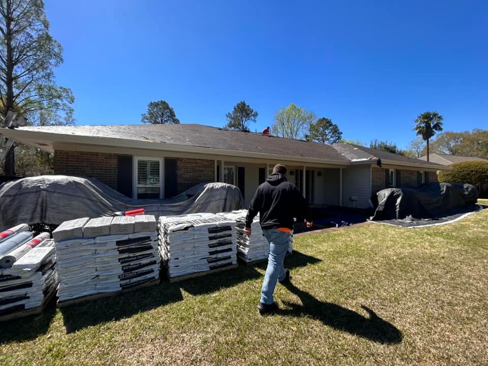A roofer walking towards a house with new shingles. Pallets of shingles sit in the foreground. The sky is blue.