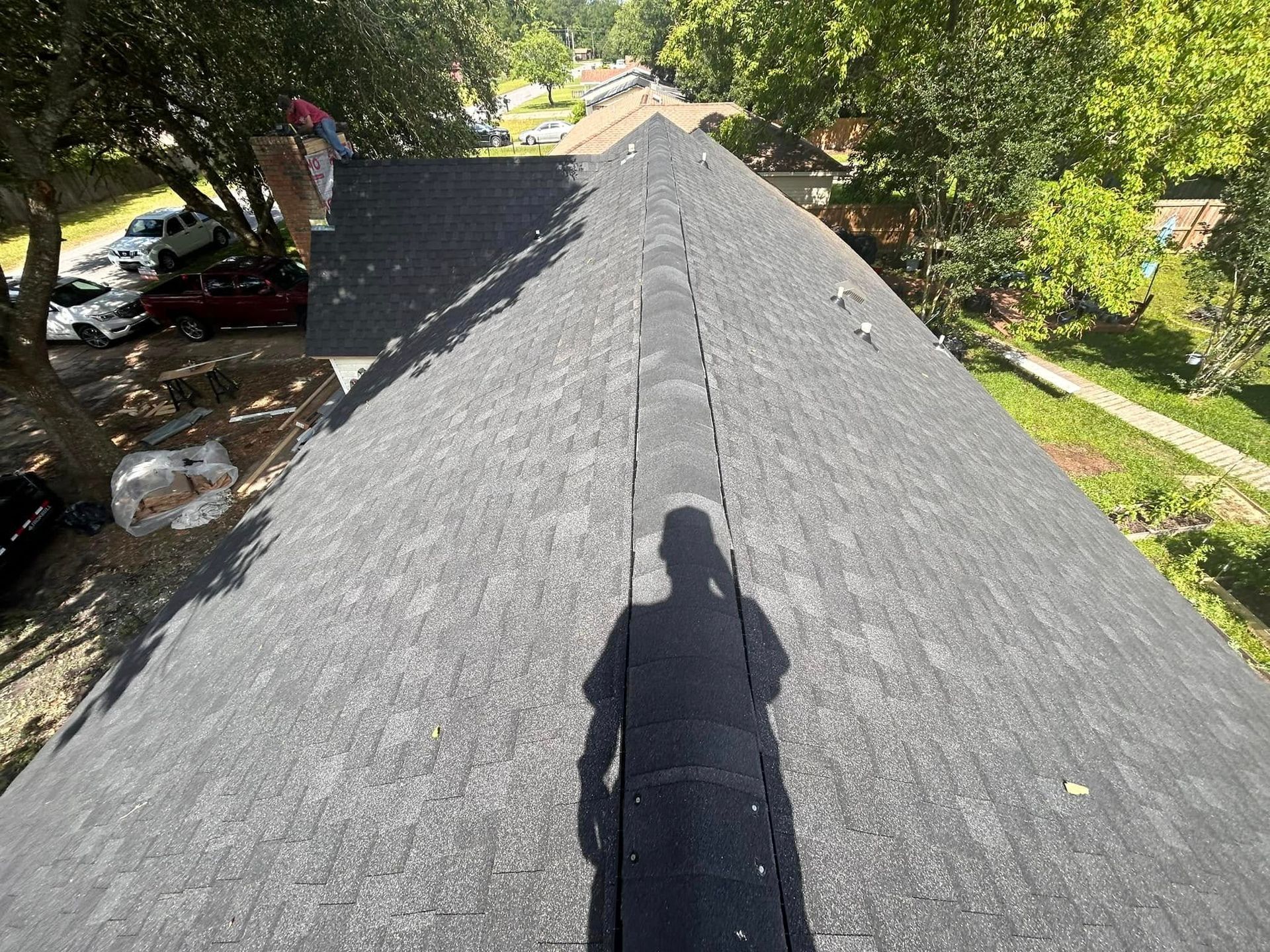 A person's shadow falls on a dark asphalt shingle roof. The roof's peak runs down the center of the frame, trees and parked cars in the background.