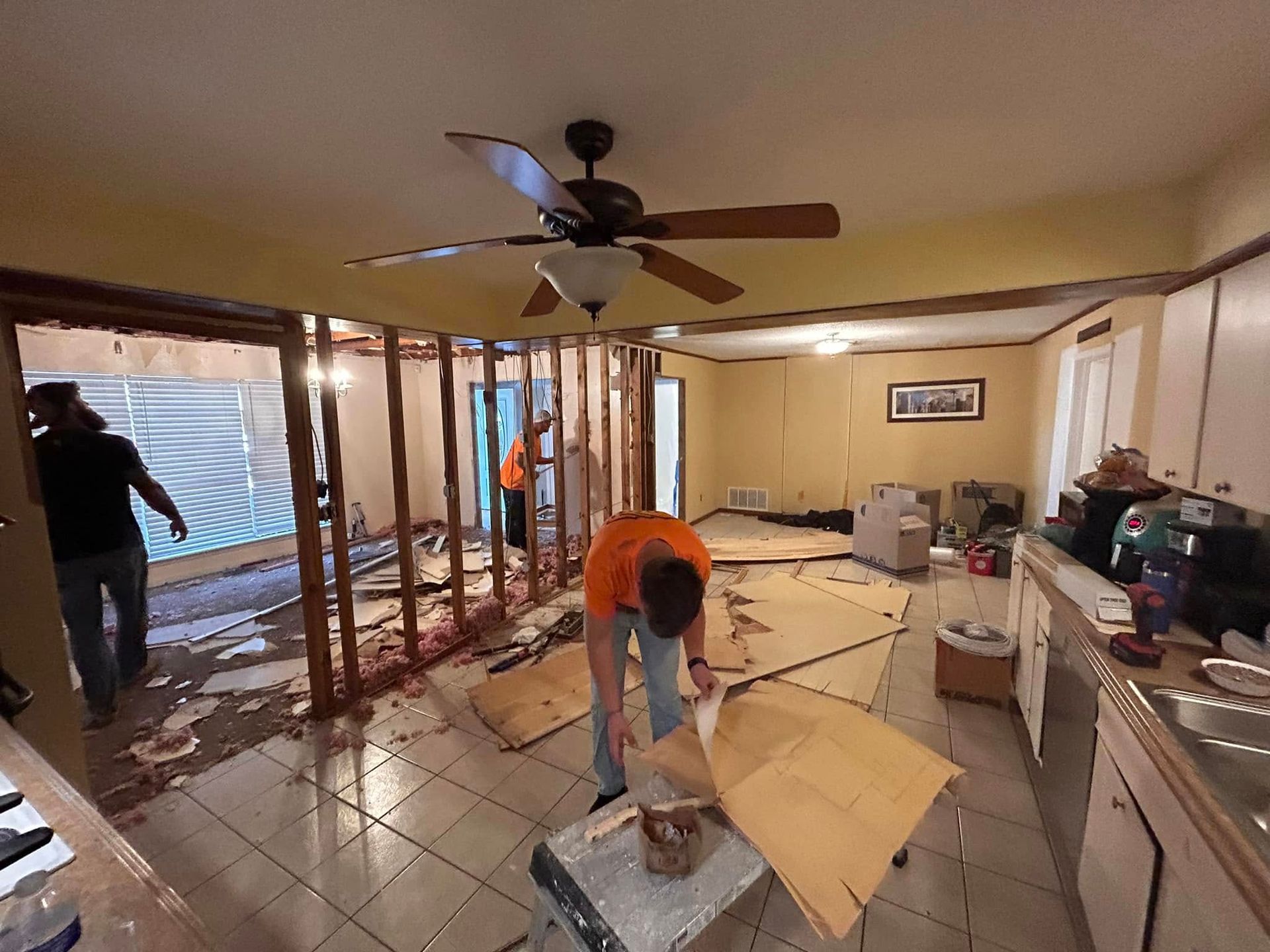 Three people are renovating a room. Drywall and debris are on the floor. One person cuts cardboard while others work on exposed wall framing.