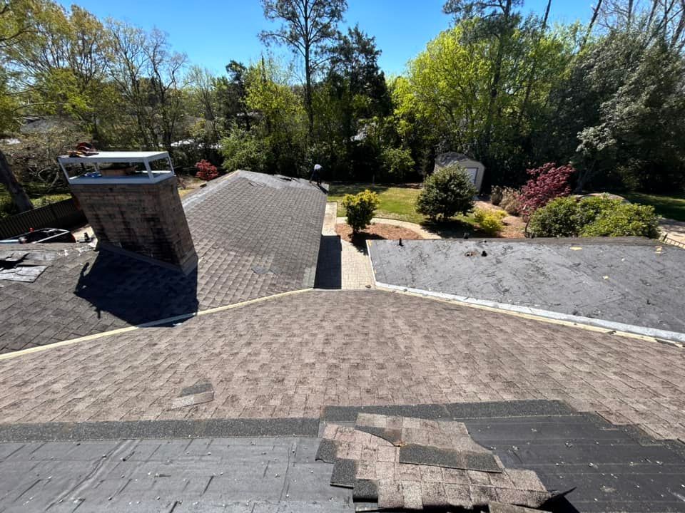 A roof under repair with exposed shingles and a chimney against a backdrop of trees and a blue sky.