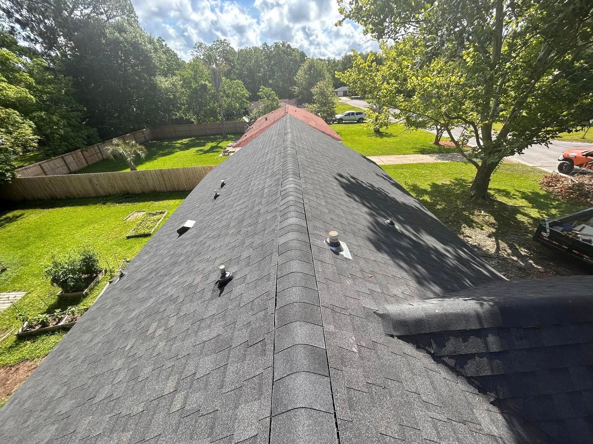 View of a dark-shingled roof, with the ridge line in the center. Green trees and grass are in the background under a sunny sky.