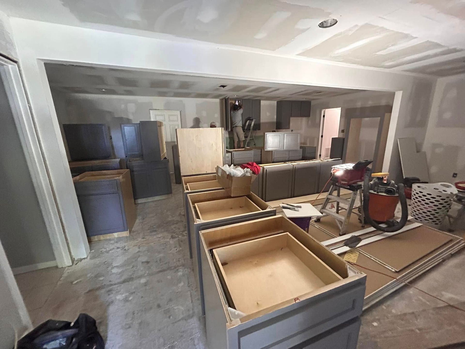 Kitchen undergoing renovation with gray cabinets, open cabinetry boxes, and exposed construction materials. Drywall and unfinished walls visible.