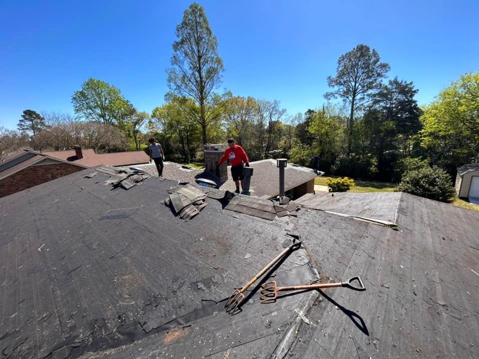 Two people stand on a dark roof, removing old shingles. Shovels and debris are scattered, trees and a blue sky in the background.