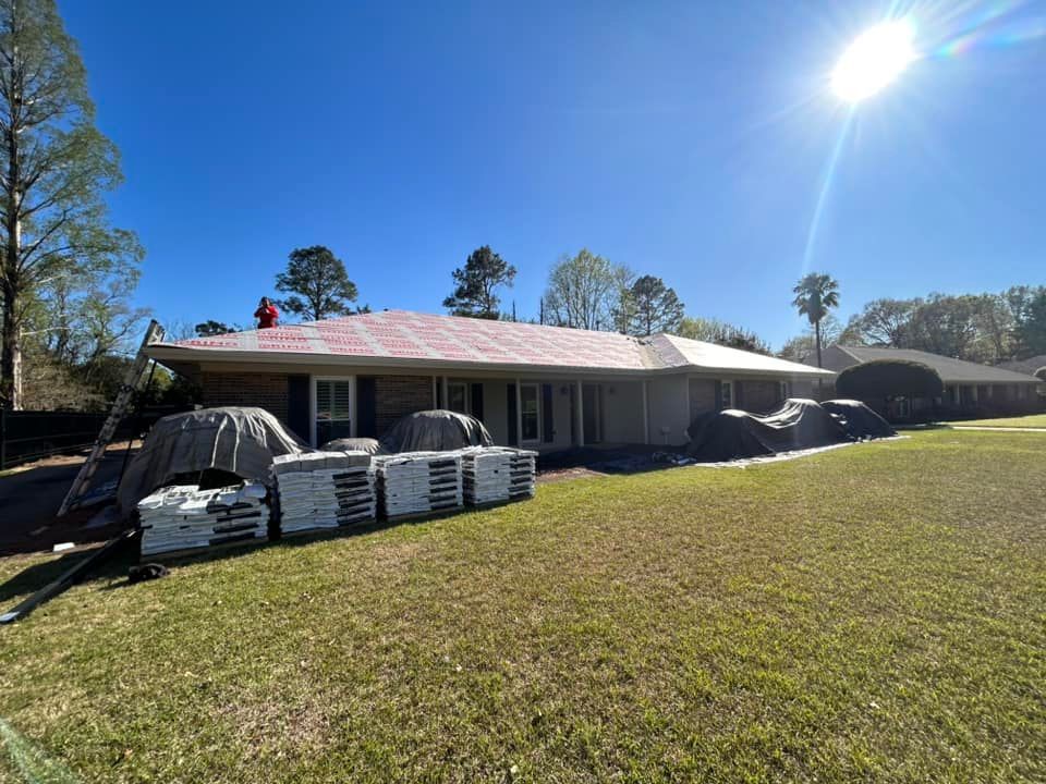 Roofers working on a residential roof under a bright sun. Pallets of roofing materials are in the foreground, and tarps cover objects around the house.