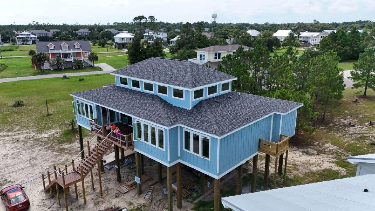 A blue stilted beach house with a gray roof and multiple windows, located in a coastal neighborhood. A wooden staircase leads to the porch.
